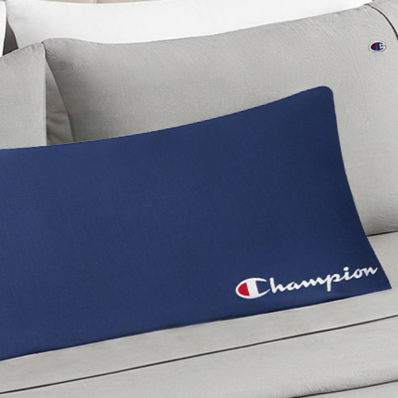 A blue Champion Jersey Body Pillow featuring white text and the brand’s signature red, white, and blue logo rests atop a light gray Champion bedding set, with both showcasing the iconic logo for a sporty touch.