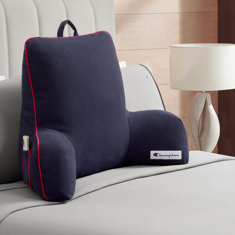 A dark gray Champion Faux Wool Backrest Pillow with red piping rests on a neatly made bed. Made from soft faux wool, the pillow offers supportive arms, a convenient side pocket, and sports a Champion label. A lamp and part of a nightstand can be seen next to the bed.