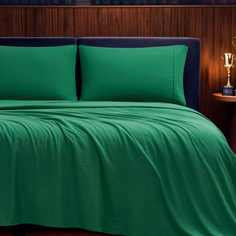 A neatly made bed features the green Champion Elastic Logo Microfiber Sheet Set and pillows, complemented by a dark headboard, a wood-paneled wall, and a small bedside table displaying a golden trophy.