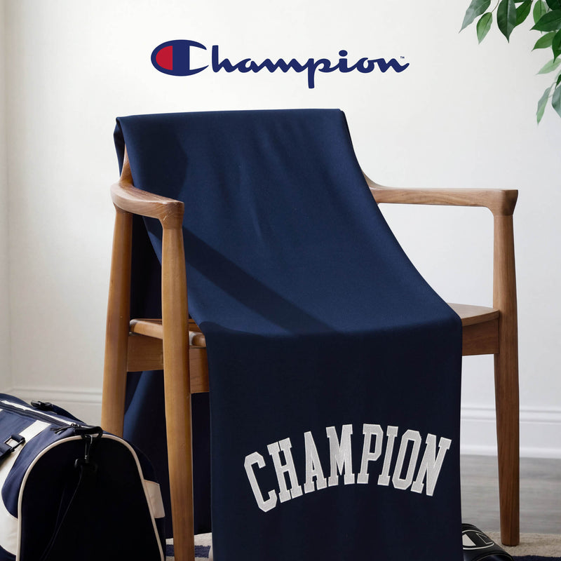 A wooden chair is draped with the Champion Jersey with Fleece Backing Throw Blanket in navy blue, its soft fleece material featuring the iconic Champion logo prominently above. Nearby, a navy and white duffel bag complements the cozy, athletic atmosphere of the scene.