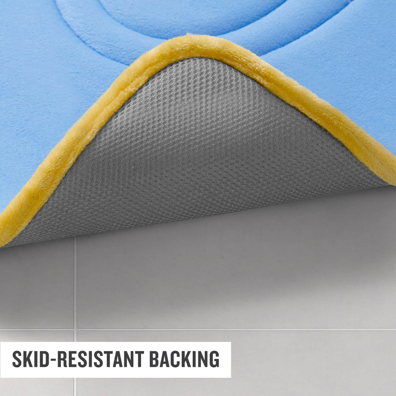 A corner of a blue and yellow Champion Quilted Logo Memory Foam Bath Mat is lifted to reveal its gray skid-resistant backing on a tiled floor. Text reads: SKID-RESISTANT BACKING.
