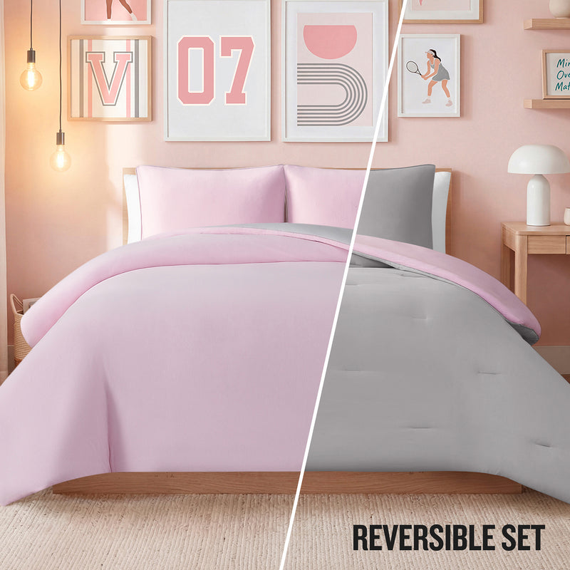 A bedroom styled with Champion’s Reversible Solid Jersey Comforter Set—cozy bedding that features pink on one side and gray on the other. Modern wall art, ambient lighting, and a compact desk complete the look. Text reads REVERSIBLE SET.