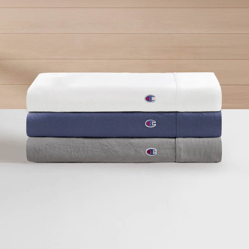 Three folded bedsheets from the Champion Embroidery Patch Microfiber Sheet Set in white, blue, and gray are stacked on a white surface. Each sheet displays a small embroidered Champion patch logo. The background consists of light wood paneling.