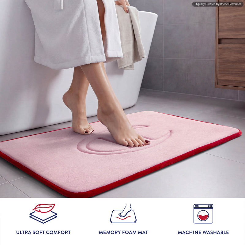 A person steps onto a pink Champion Quilted Logo Memory Foam Bath Mat featuring plush memory foam in a bathroom. Icons below highlight features: ultra soft comfort, memory foam, and machine washable.