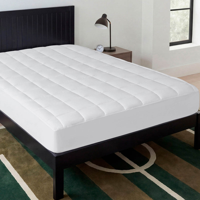 A neatly made bed features the Champion Ultimate Protection Mattress Pad, a white, water-resistant topper, in a modern bedroom with a black headboard, bedside table with lamp and clock, and a geometric-patterned rug on the floor.
