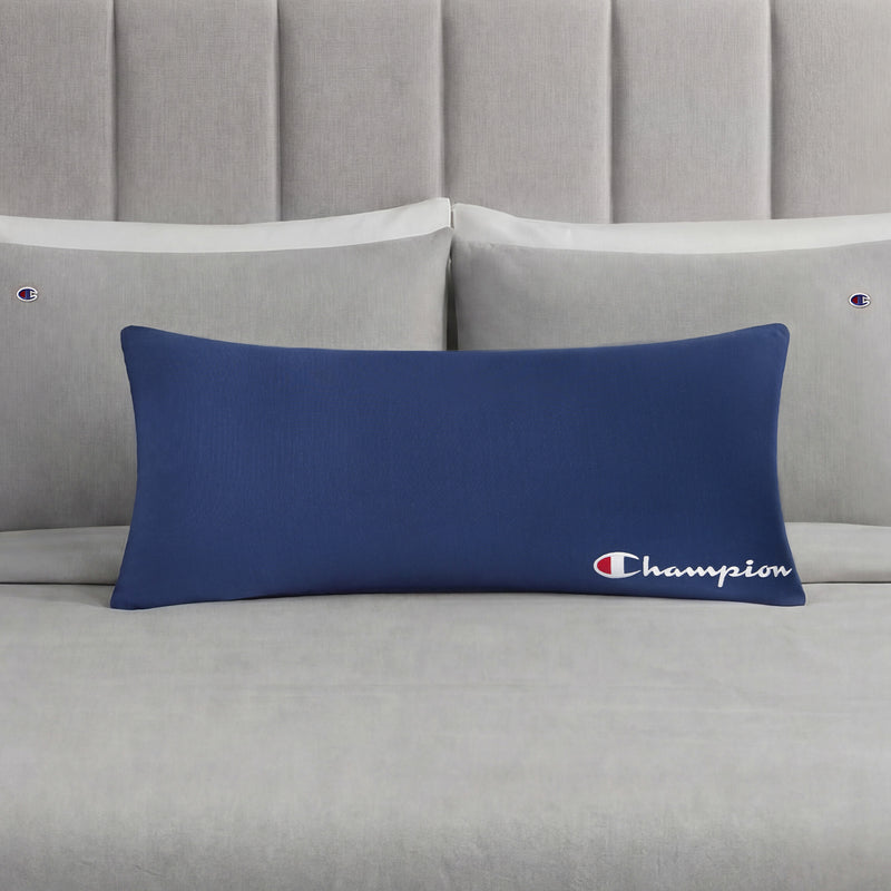 A Champion Jersey Body Pillow in bold blue rests on a neatly made bed with light gray Champion-branded pillows and sheets. A light gray tufted headboard enhances the understated sporty vibe of the room.