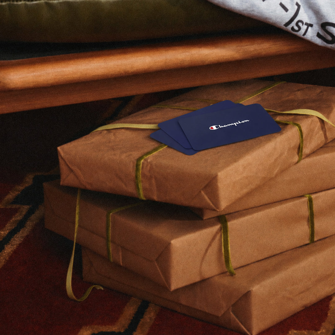 Three wrapped gift boxes in brown paper with olive green ribbons are stacked on a patterned rug under a wooden bench. A blue Champion card is placed on top of the boxes.