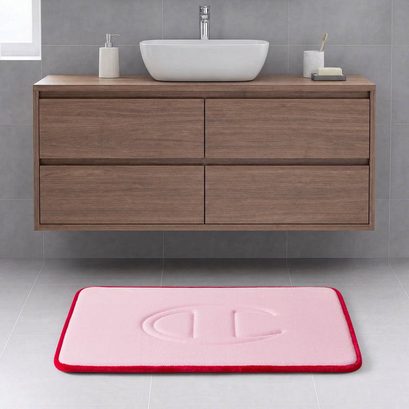 A modern bathroom with a wooden floating vanity, a white rectangular basin, and the Quilted Logo Memory Foam Bath Mat by Champion—featuring a pink surface with a red border and an embossed design—placed on a light gray tiled floor.