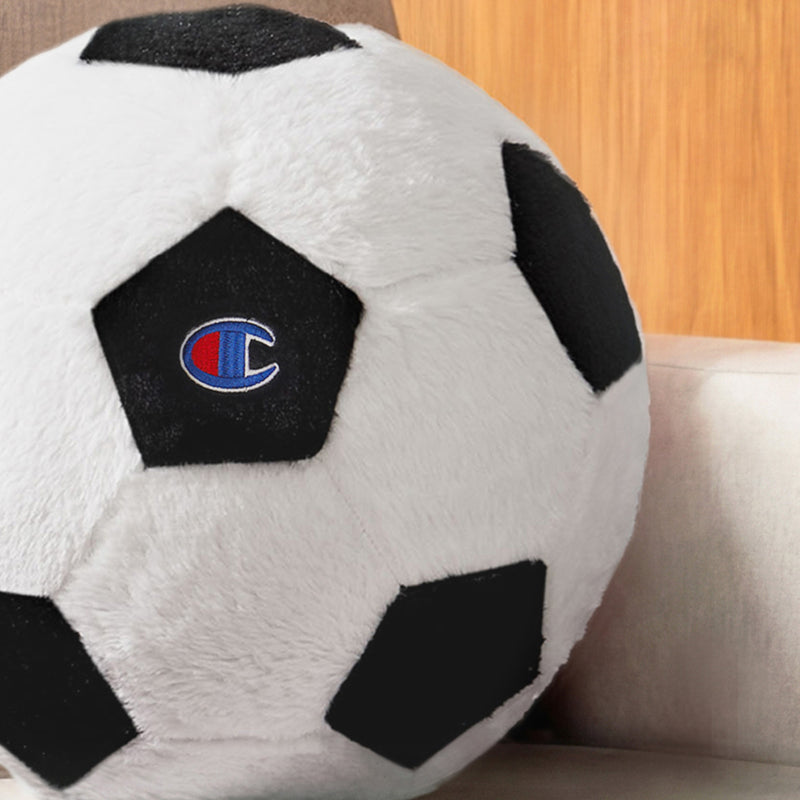 A cozy corduroy Champion Sport Shaped Pillow, designed like a black and white football and featuring an embroidered blue and red Champion logo on one panel, rests on a light-colored couch. A wooden wall in the background adds warm contrast to this decorative accent.