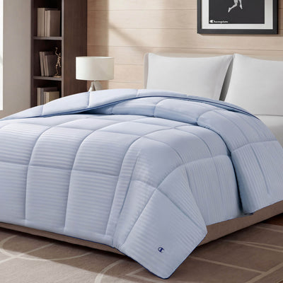 A Blue Cooling Down Alternative Comforter by Champion is displayed on a bed in a modern bedroom with white pillows, a lamp, bookshelf, and framed picture on the wall. The space features neutral decor and soft lighting.