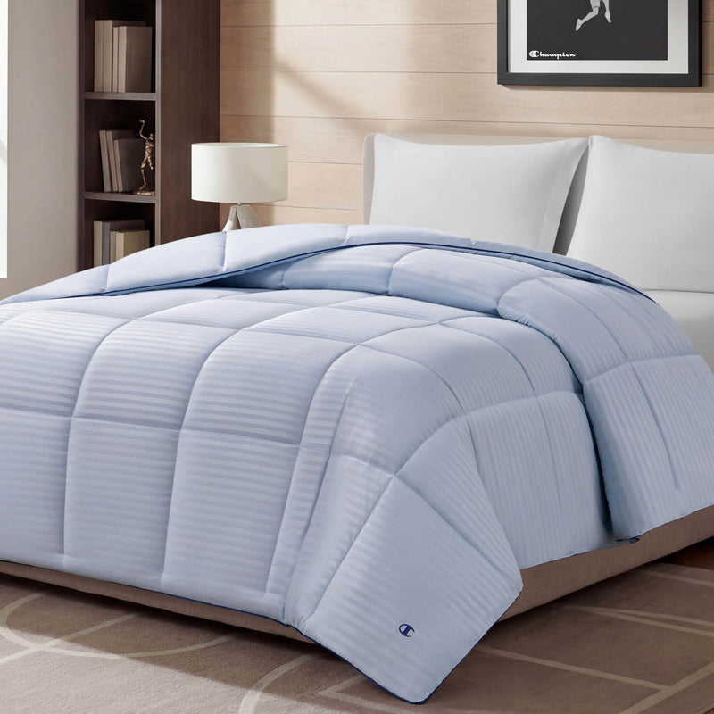 A Blue Cooling Down Alternative Comforter by Champion is displayed on a bed in a modern bedroom with white pillows, a lamp, bookshelf, and framed picture on the wall. The space features neutral decor and soft lighting.