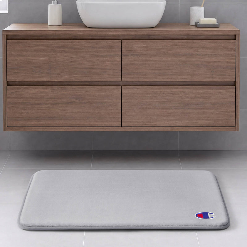 A light gray Champion Logo Patch Memory Foam Bath Mat with a small emblem in the corner rests on a light gray tiled floor in front of a floating wooden bathroom vanity.