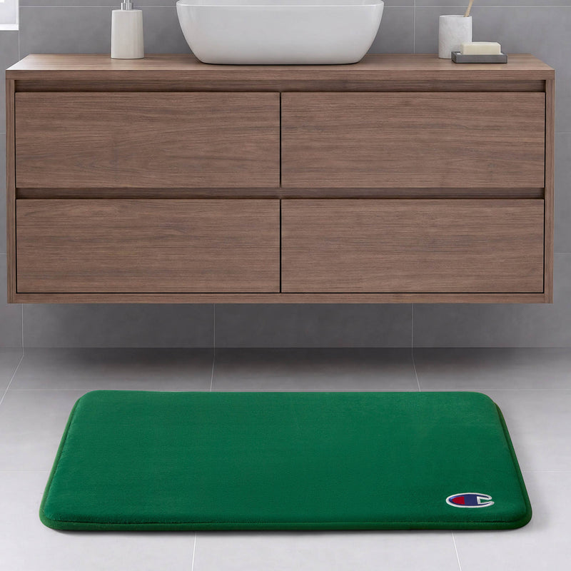 A Champion Logo Patch Memory Foam Bath Mat in green, featuring a small logo in the corner, rests on a light gray bathroom floor in front of a modern wooden vanity with four drawers. Designed by Champion, this skid-resistant bath mat is crafted from memory foam to provide superior comfort and safety during your daily routine.