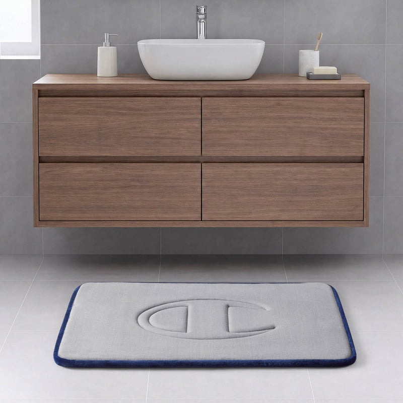 A modern bathroom with a floating wooden vanity, a vessel sink, and a rectangular gray Quilted Logo Memory Foam Bath Mat from Champion, featuring an embossed logo, set on a light tile floor.