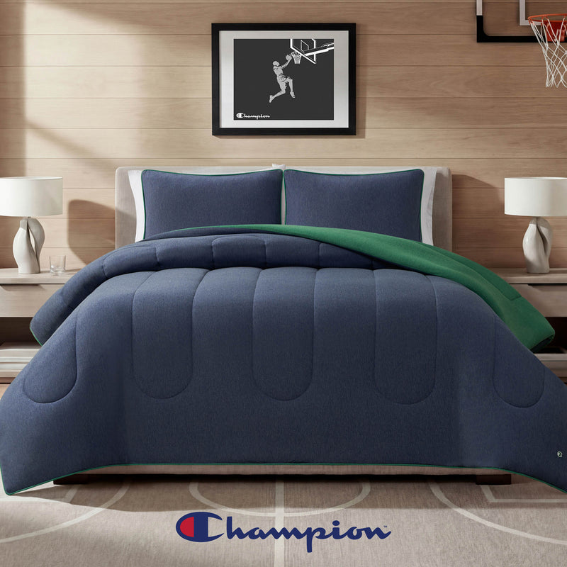 A neatly made bed features the Champion Premium Reversible Solid Jersey Comforter Set in blue and green, complemented by two blue pillows and a Champion logo displayed at the foot. Two lamps rest on bedside tables, while a framed basketball art piece hangs above the bed.