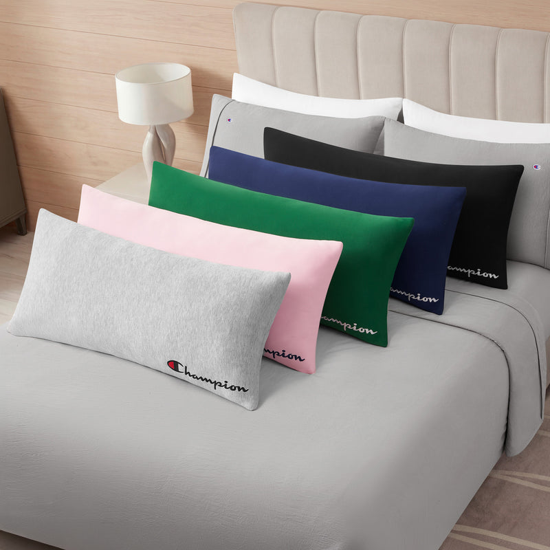 Five Champion Jersey Body Pillows—including athletic accents in gray, pink, green, navy blue, and black—are neatly arranged on a modern bed dressed in gray bedding. Each jersey knit body pillow adds comfort and sporty style to the room, while a lamp rests on the nightstand beside the bed.