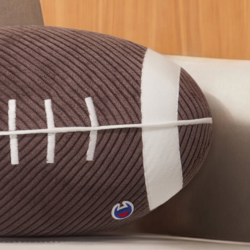 A Champion Sport Shaped Pillow, designed like an American football with white laces and stripes, rests on a beige couch.