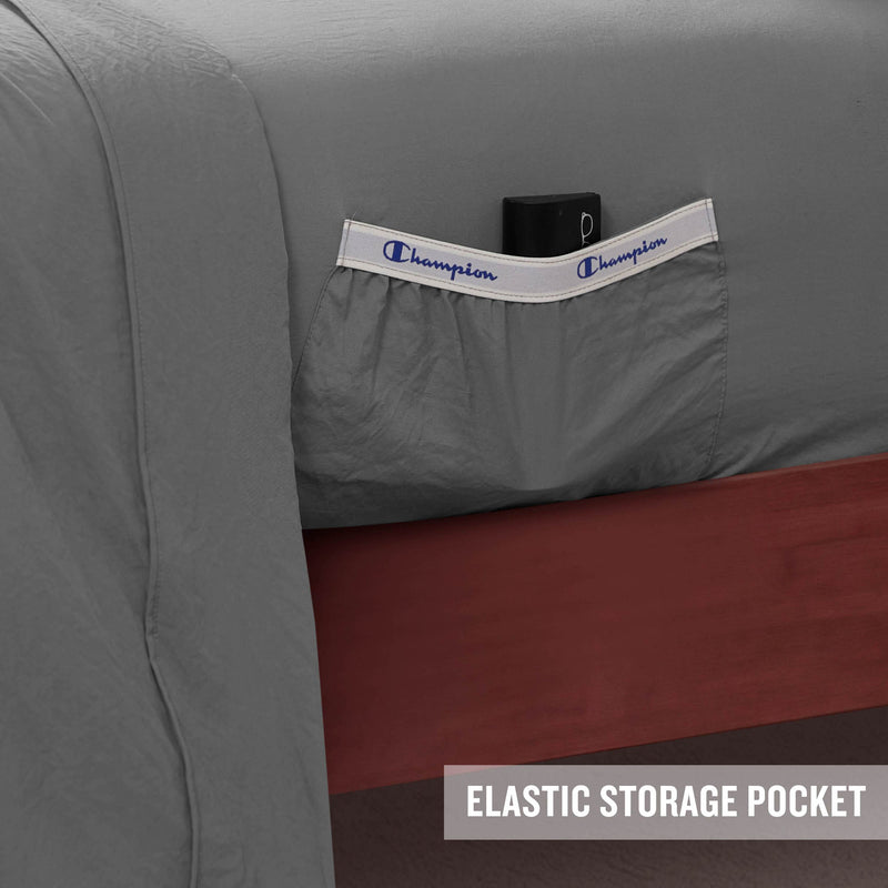 A gray Peak Comfort Microfiber Sheet Set by Champion features an elastic Champion-branded side pocket holding a black item. The sheets are displayed on a wooden bed frame, with an “ELASTIC STORAGE POCKET” label in the corner highlighting the practical design of these deep pocket Champion sheets—ideal for anyone seeking athletic bedding with convenient storage solutions.
