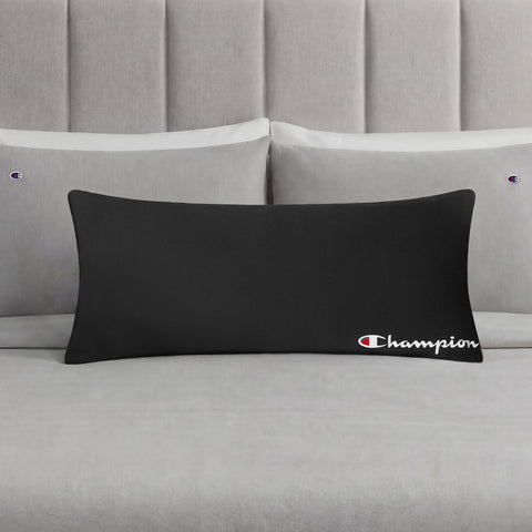 A Champion Jersey Body Pillow in black is displayed on a neatly made bed with light gray bedding and matching pillowcases, each embroidered with small Champion logos, making it the perfect athletic accent pillow for any modern bedroom.