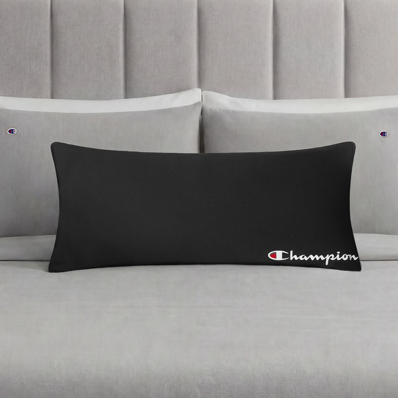 A Champion Jersey Body Pillow in black is displayed on a neatly made bed with light gray bedding and matching pillowcases, each embroidered with small Champion logos, making it the perfect athletic accent pillow for any modern bedroom.