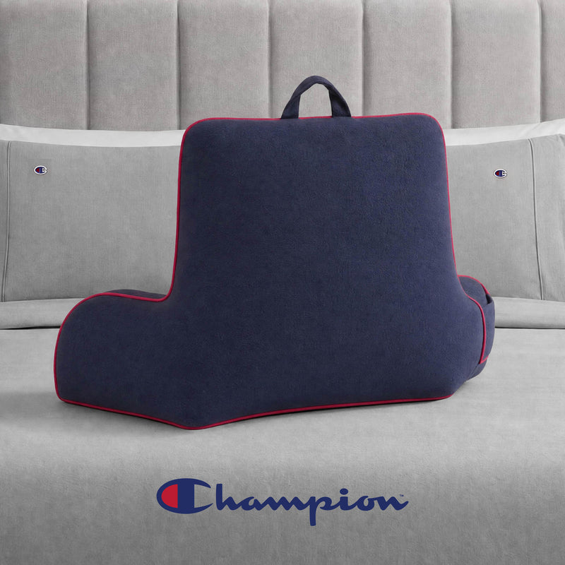 A Champion Faux Wool Backrest Pillow in navy blue with red trim and a cozy faux wool finish sits on a gray bed, complementing the Champion branding on the pillows and at the foot of the bed.
