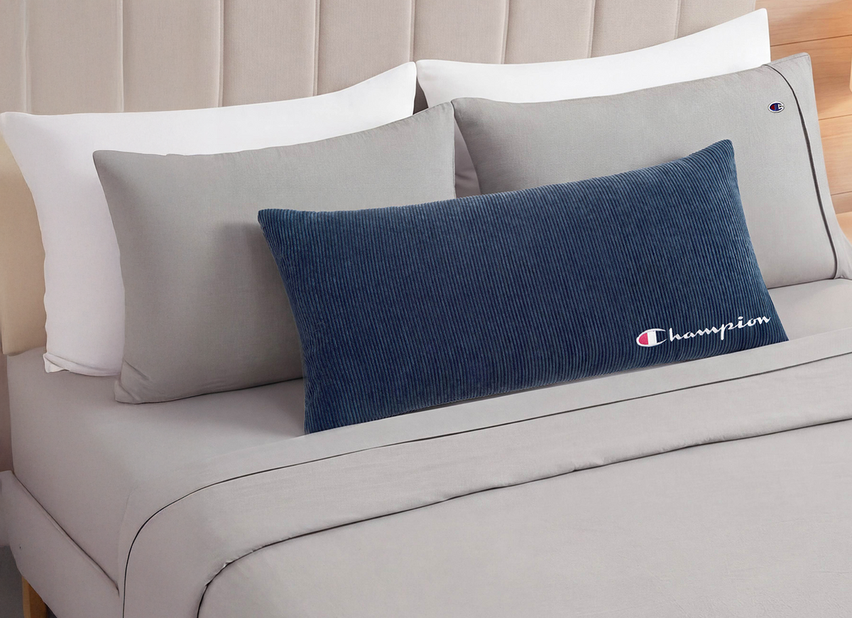 A navy blue corduroy lumbar pillow with an embroidered Champion logo, styled on a bed with light gray and white pillows against a beige headboard.