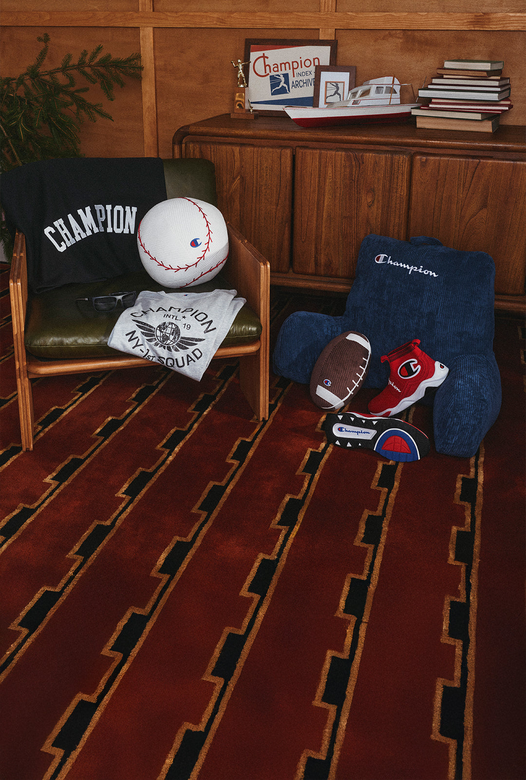 A styled interior scene with two wooden chairs and a matching rug. The left chair has a green seat and holds a black Champion sweatshirt, a baseball, and a white Champion T-shirt.