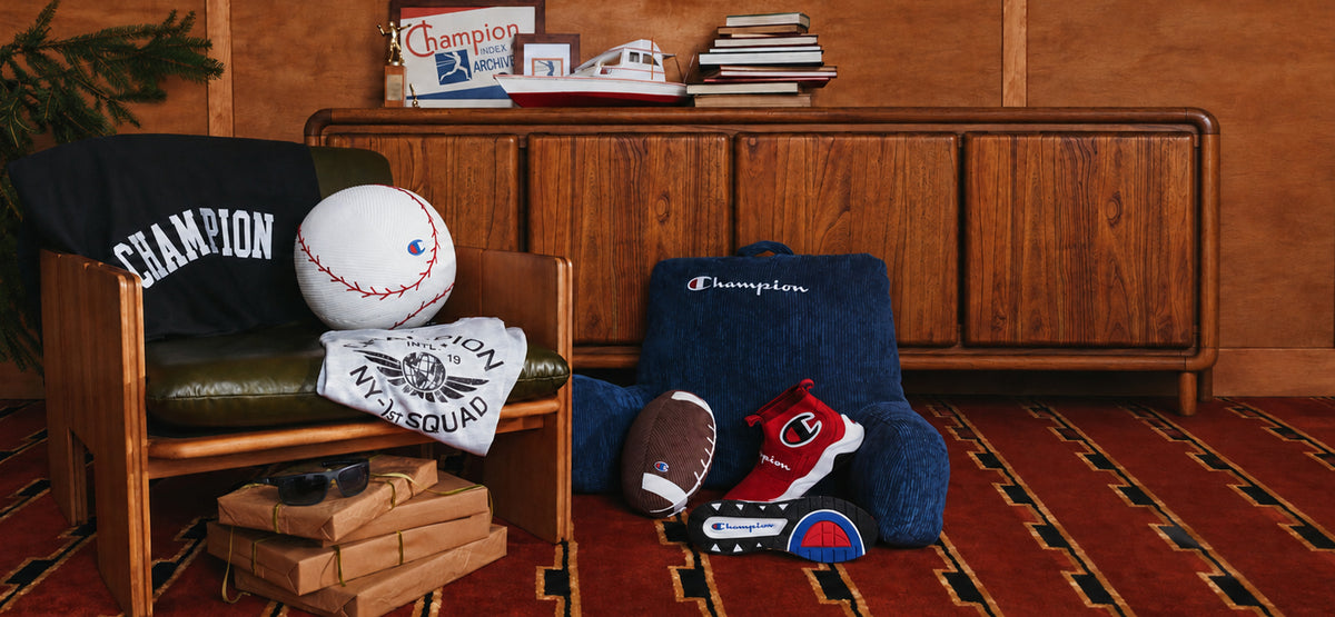 A styled interior scene with multiple Champion-branded sports-themed products arranged in a room: a black Champion chair, a white baseball, a white towel with a team logo, a navy Champion headrest pillow, a football, a red-and-blue shoe, and a small toy car on a patterned rug in front of a wooden cabinet.