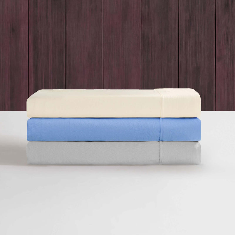 Three neatly folded Champion Elastic Logo Microfiber Sheet Sets, in cream, blue, and gray, are stacked on a white surface against a dark wood-paneled background—offering everyday comfort and featuring subtle elastic logo detail.