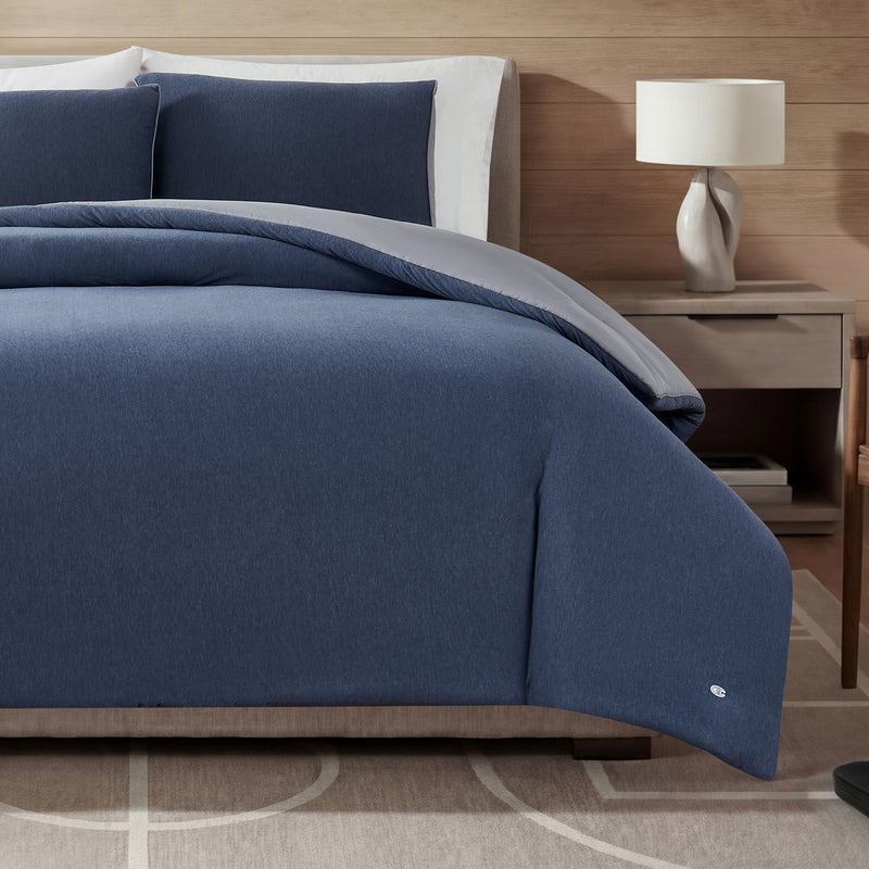 A neatly made bed features the Champion Reversible Solid Jersey Comforter Set in blue, with matching pillows arranged against a wooden headboard. On the right side, a white lamp rests on a nightstand, while a patterned beige rug covers the floor. The reversible comforter set adds both style and versatility to the room’s décor.