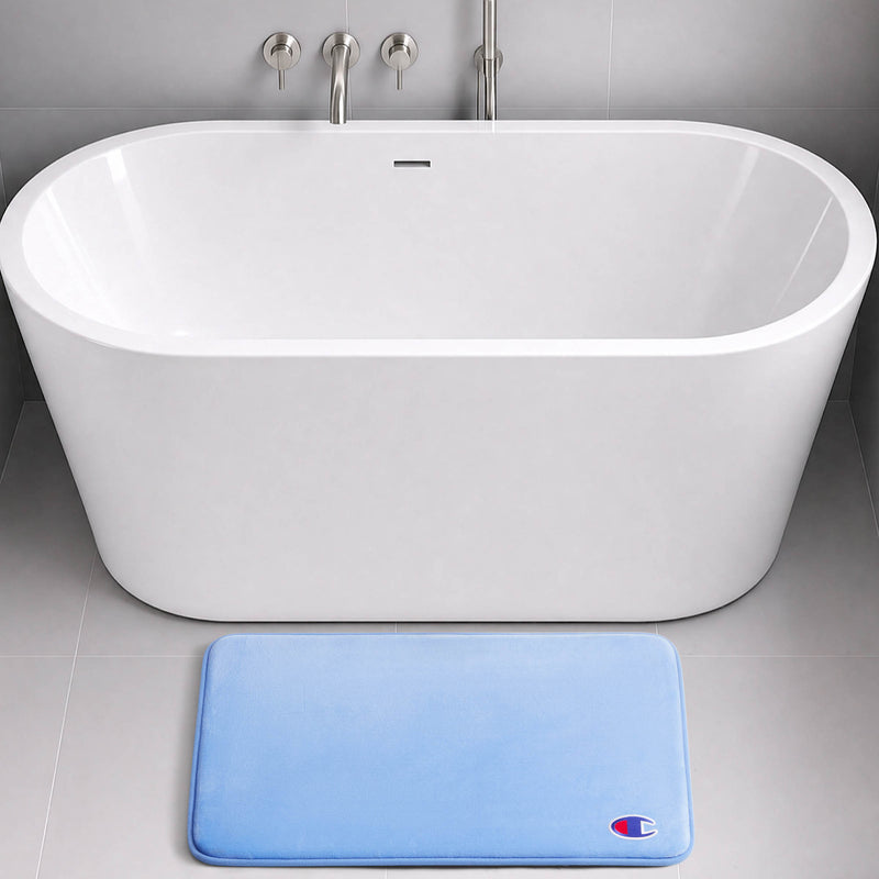 A modern white freestanding bathtub with wall-mounted fixtures sits on a gray tile floor. In front of the tub is a rectangular light blue Logo Patch Memory Foam Bath Mat from Champion, featuring a small Champion logo patch in the bottom right corner. The Champion bath mat offers added comfort and support with its plush memory foam construction.