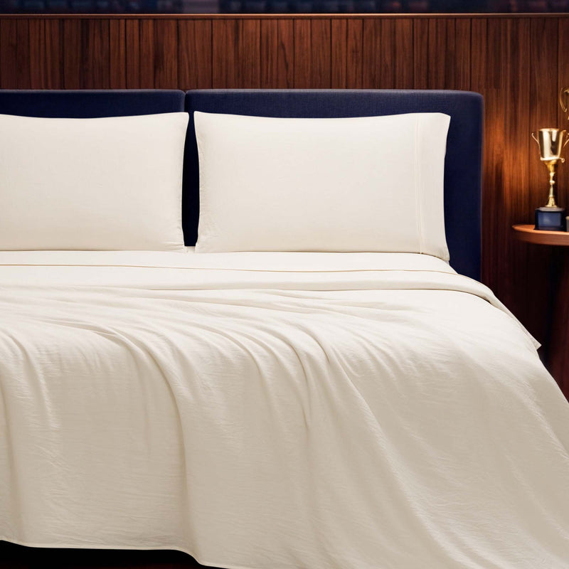 A neatly made bed with white sheets and pillows from the Champion Elastic Logo Microfiber Sheet Set exudes everyday comfort, complemented by a blue headboard and a wood-paneled wall. A small table with a golden trophy is visible on the right side, while an elastic logo detail on the bedding adds a touch of modern style.