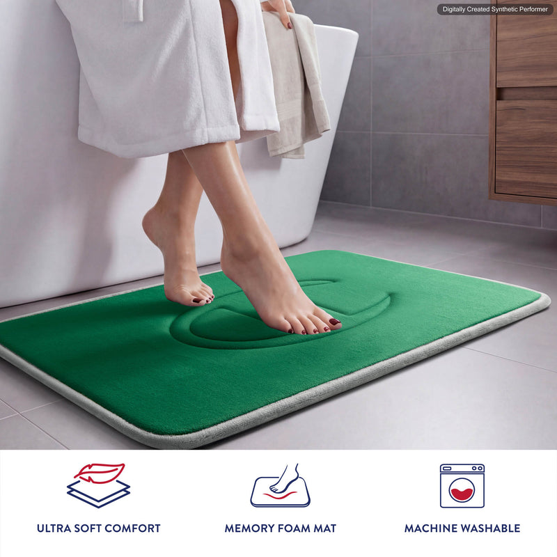 A person in a white robe steps onto a green Champion Quilted Logo Memory Foam Bath Mat next to a bathtub. Icons below highlight its ultra-soft comfort, supportive memory foam, and machine-washable design.