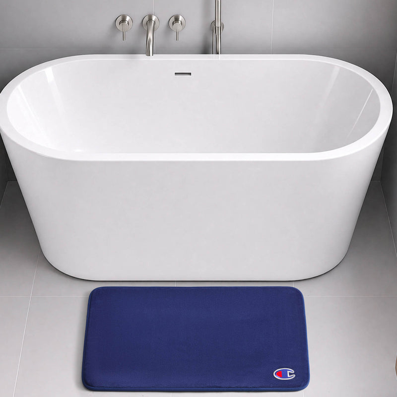 A modern white freestanding bathtub with chrome fixtures sits on a gray tiled floor. In front of the tub, a dark blue Champion Logo Patch Memory Foam Bath Mat with skid-resistant backing and a small logo offers both style and safety.