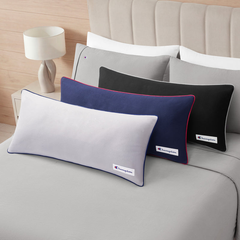 Three rectangular Champion Faux Wool Body Pillows—in gray, navy blue, and black—featuring the iconic Champion logo are stacked on a neatly made bed with gray bedding. Each pillow boasts a plush polyester fill for extra comfort. A lamp sits on a nightstand next to the bed.