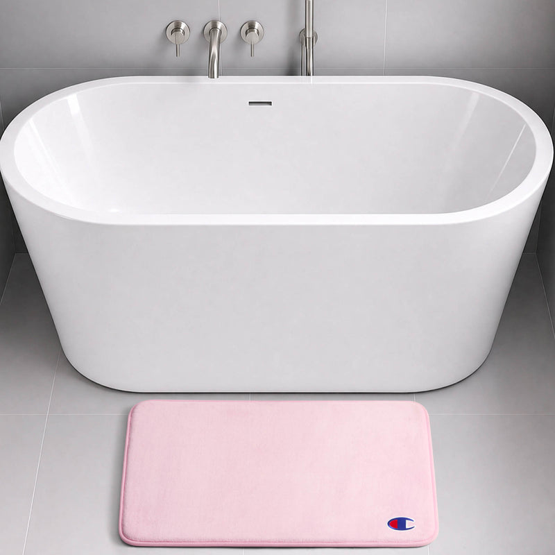 A modern white freestanding bathtub with silver fixtures is set against a gray tiled wall. On the floor in front of the tub lies a light pink rectangular Logo Patch Memory Foam Bath Mat by Champion, featuring a skid-resistant design and adorned with a small blue and red logo.