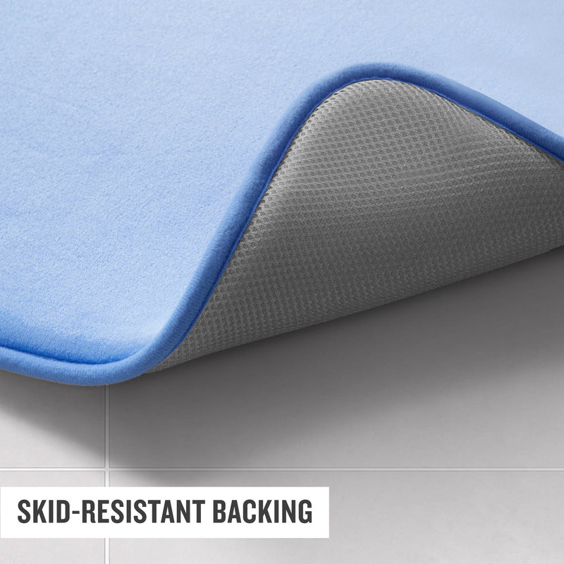 A close-up of a blue Champion Logo Patch Memory Foam Bath Mat on a tiled floor, with one corner flipped to reveal its gray, textured, skid-resistant backing. Text reads: Skid-resistant backing.