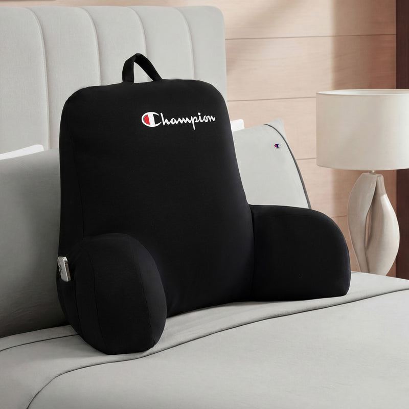 A black Champion Jersey Backrest Pillow with arms rests on a neatly made bed, the iconic Champion logo visible, beside a light-colored lamp on the nightstand in a modern bedroom.