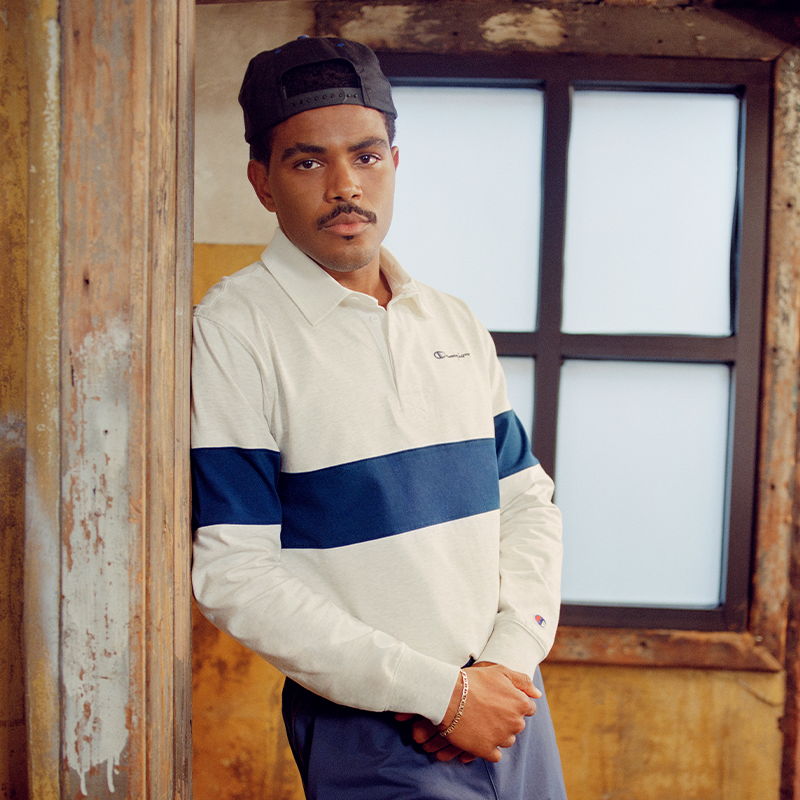 A man wearing a white long-sleeve polo shirt with a navy blue chest stripe and a small Champion logo on the left chest, paired with a black cap and dark pants, leaning against a rustic wooden door frame.