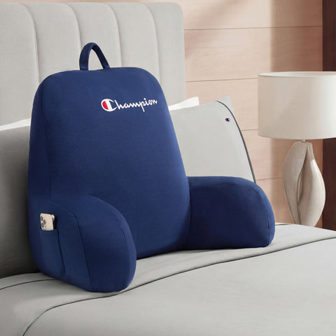 A Champion Jersey Backrest Pillow in navy blue, featuring armrests and crafted from soft jersey knit fabric, rests on a neatly made bed. A smartphone is stored in its convenient side pocket, while a lamp and nightstand appear in the background.