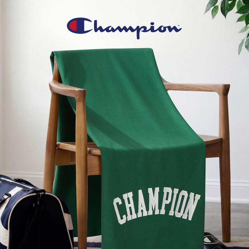 A Champion Jersey with Fleece Backing Throw Blanket in green is draped over a wooden chair, featuring the Champion logo above and a duffel bag resting on the floor nearby. This soft and stylish fleece throw brings both comfort and a sporty touch to any space.