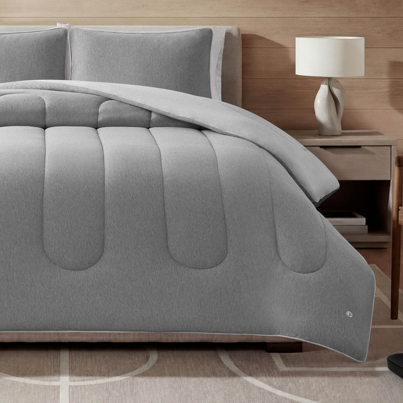 A neatly made bed features the Champion Premium Reversible Solid Jersey Comforter Set with matching pillows, complemented by a wooden nightstand topped with a white lamp, all set within a modern, minimalist bedroom design.