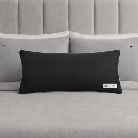 A black rectangular Champion Faux Wool Body Pillow with a small Champion logo rests on a neatly made bed with light gray sheets and two matching pillows, each displaying the Champion brand. The pillow’s plush polyester fill enhances the stylish bedding ensemble with inviting comfort.