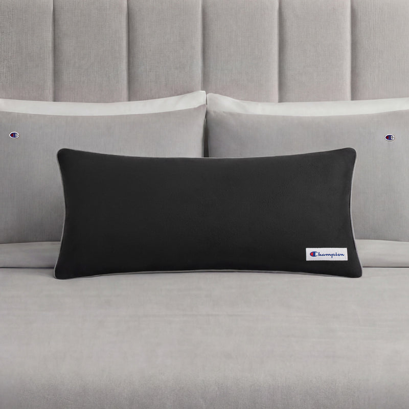 A black rectangular Champion Faux Wool Body Pillow with a small Champion logo rests on a neatly made bed with light gray sheets and two matching pillows, each displaying the Champion brand. The pillow’s plush polyester fill enhances the stylish bedding ensemble with inviting comfort.