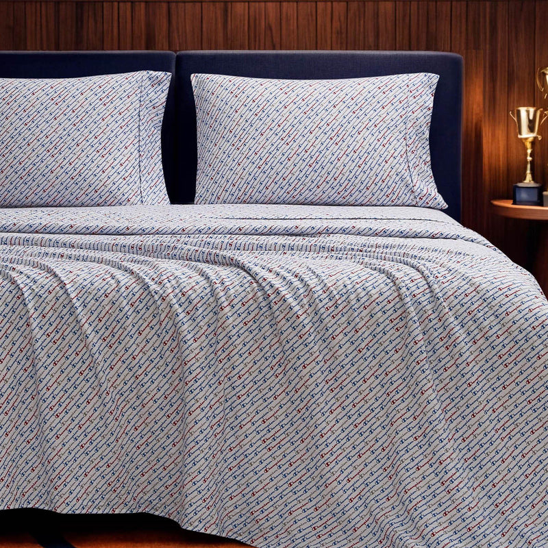 A neatly made bed features the Champion Printed Microfiber Sheet Set in blue and white patterns, paired with two matching pillows. A dark headboard stands against a wood-paneled wall, beside a small table displaying a gold trophy.