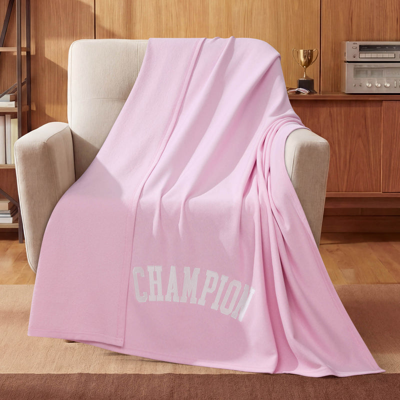 A light pink Champion Jersey with Fleece Backing Throw Blanket featuring “CHAMPION” in white letters is draped over a beige armchair. In the background, a wooden cabinet holds a trophy and a stereo system.