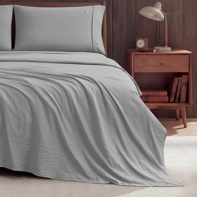 A neatly made bed with light gray Peak Comfort Microfiber Sheet Set by Champion sits next to a wooden nightstand holding a lamp, an alarm clock, and books, set against a dark wood-paneled wall on a light-colored floor.