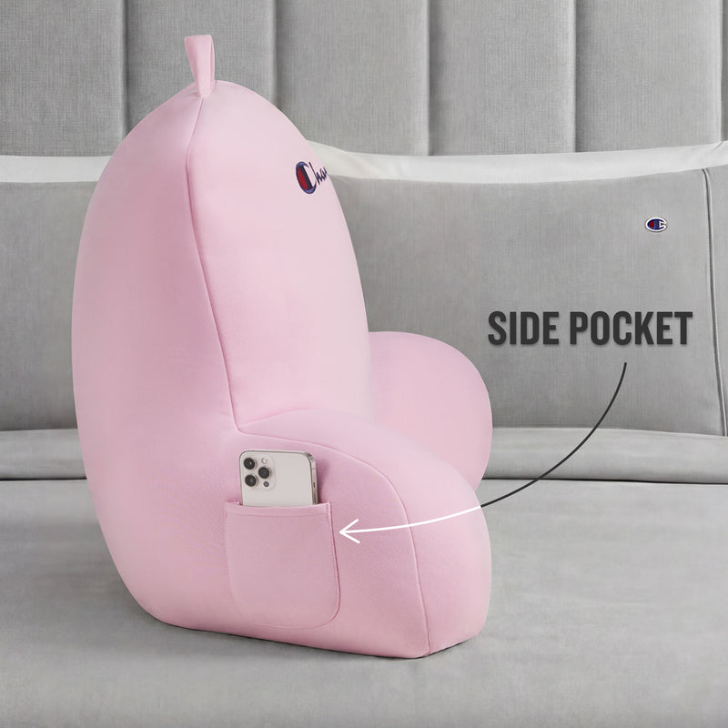 A Champion Jersey Backrest Pillow in pink rests on the bed. Made from soft jersey knit fabric, it has a handy side pocket for your smartphone—highlighted by the text “SIDE POCKET”—and is finished with an embroidered Champion logo.