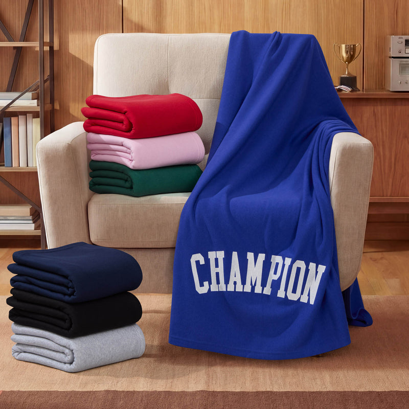A blue Champion Jersey with Fleece Backing Throw Blanket featuring bold CHAMPION text is draped over a beige armchair; nearby, folded Champion Jersey with Fleece Backing Throw Blankets in pink, red, green, navy, black, and gray are stacked together in a cozy wooden room.