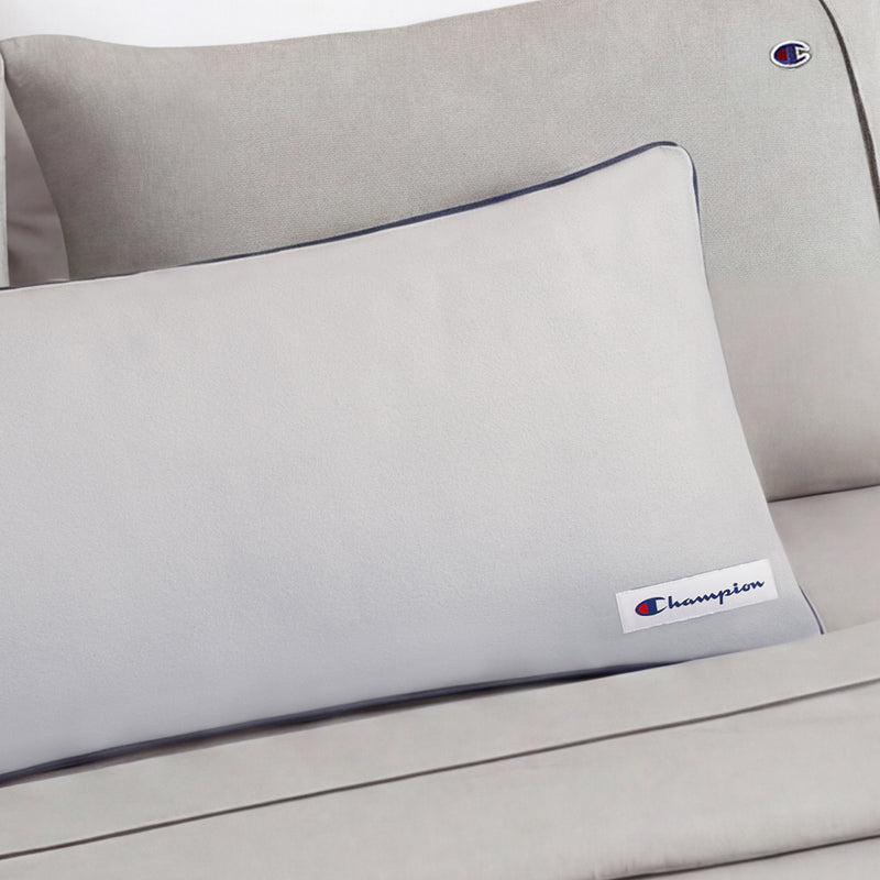 A light gray Faux Wool Body Pillow by Champion, filled with plush polyester and adorned with the Champion logo patch, is elegantly placed on a matching couch or bed for a clean, minimalistic look.